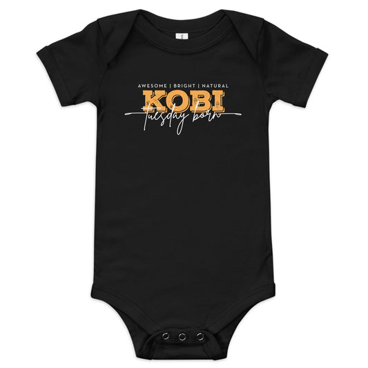 A white short-sleeve baby bodysuit with a graphic print on the chest reading 'AWESOME | BRIGHT | NATURAL KOBI tuesday born' in white and yellow lettering, shown on a plain white background